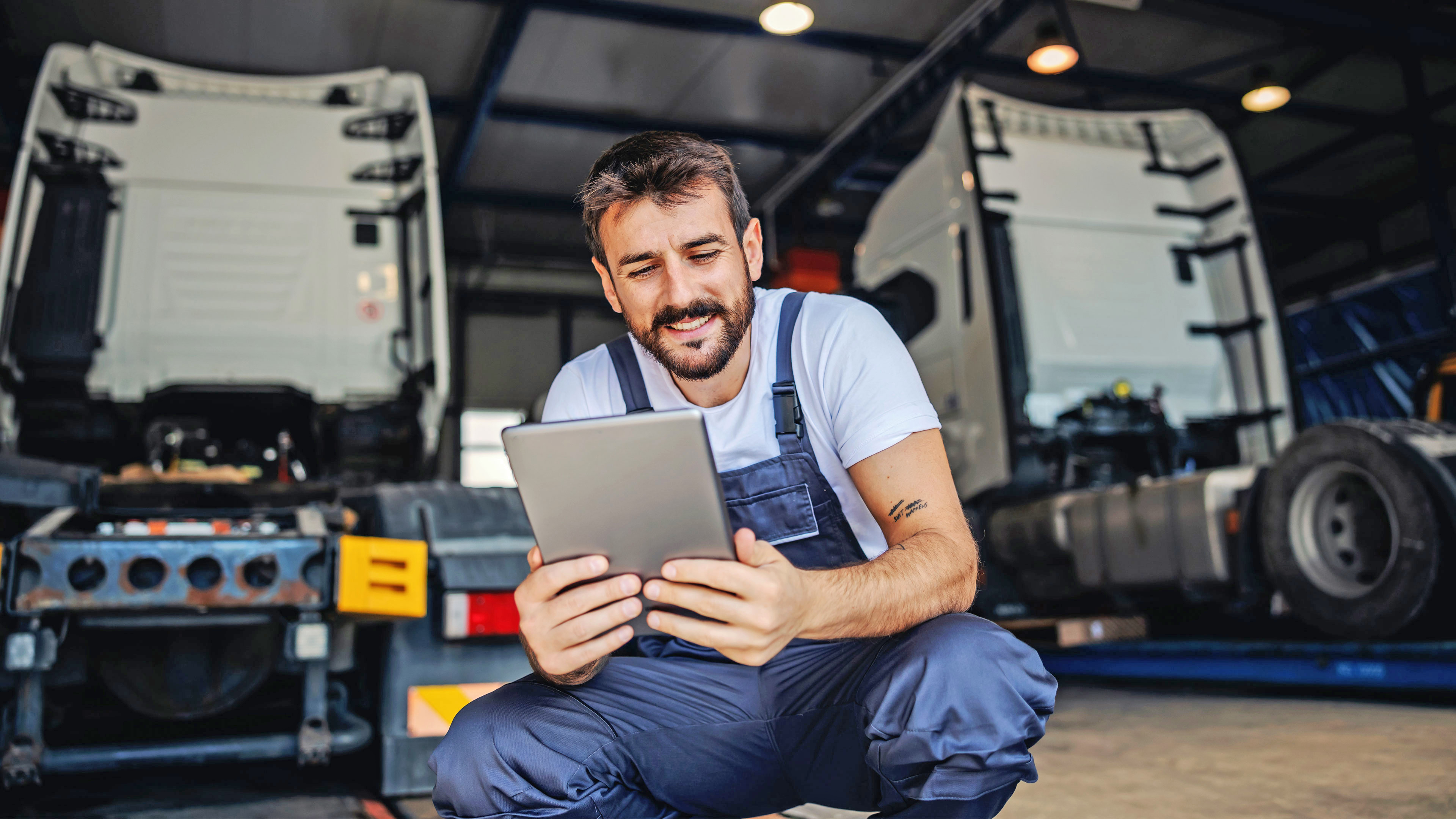 In the foreground, you can see a man in blue work clothes holding a tablet and looking at it with high interest. He is crouched down so that you can see his workshop and two trucks behind him.