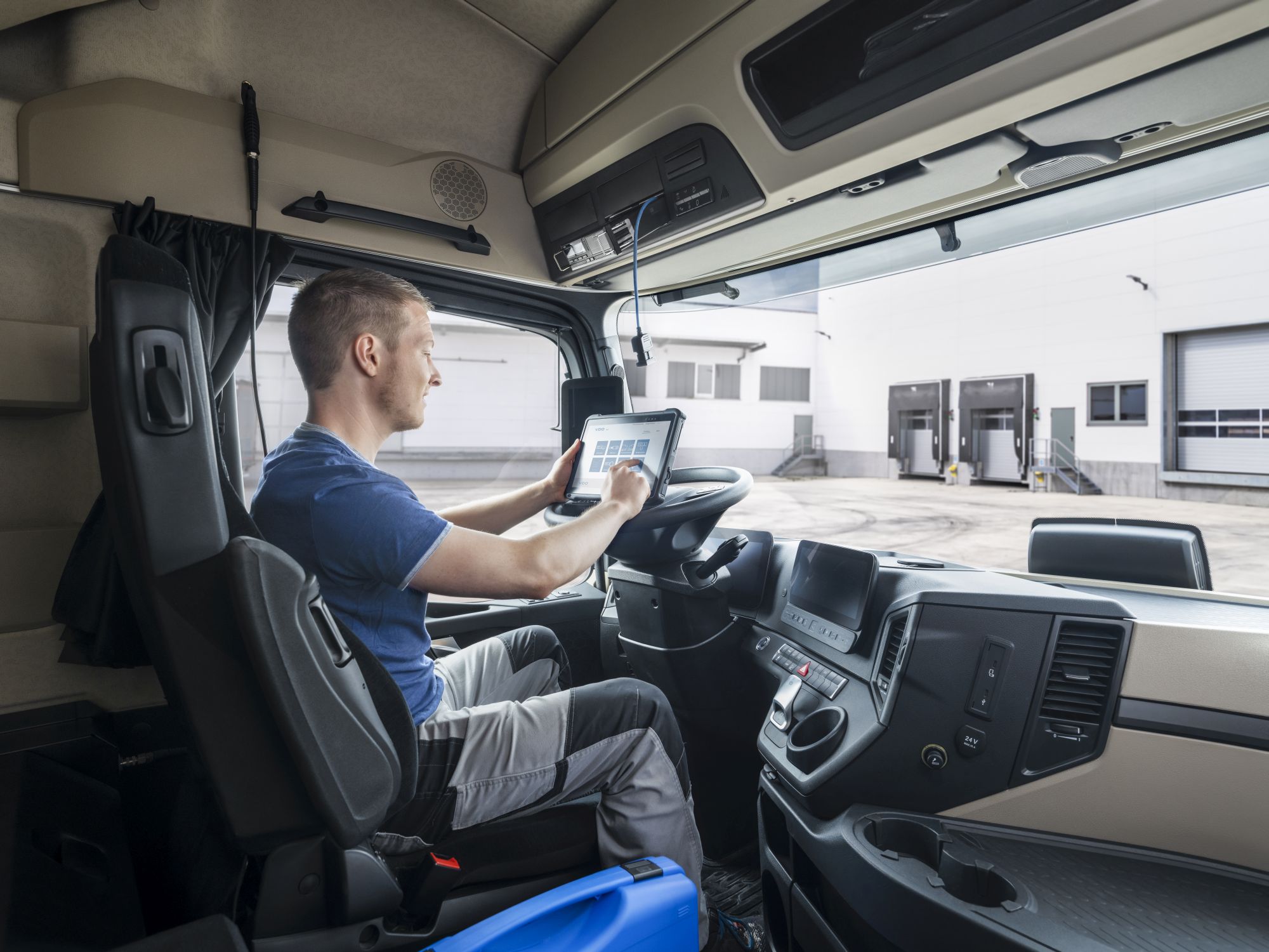 Image of a tachograph testing situation. A workshop employee is sitting in the driver's seat of a large truck, operating the VDO WorkhopTab. In the background, you see a large production hall with two loading docks.